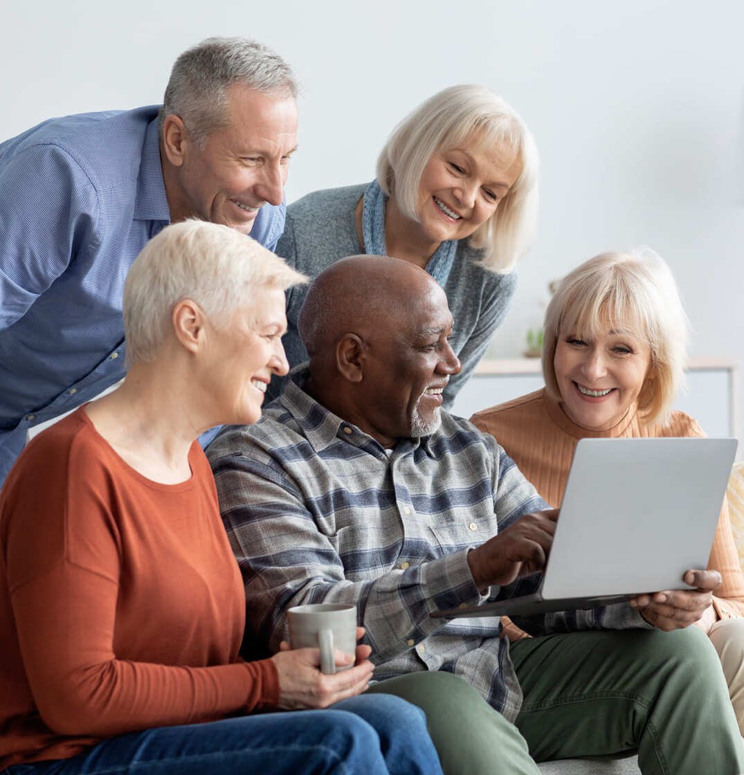 Five diverse seniors gathered around laptop computer, smiling and learning together in comfortable home setting